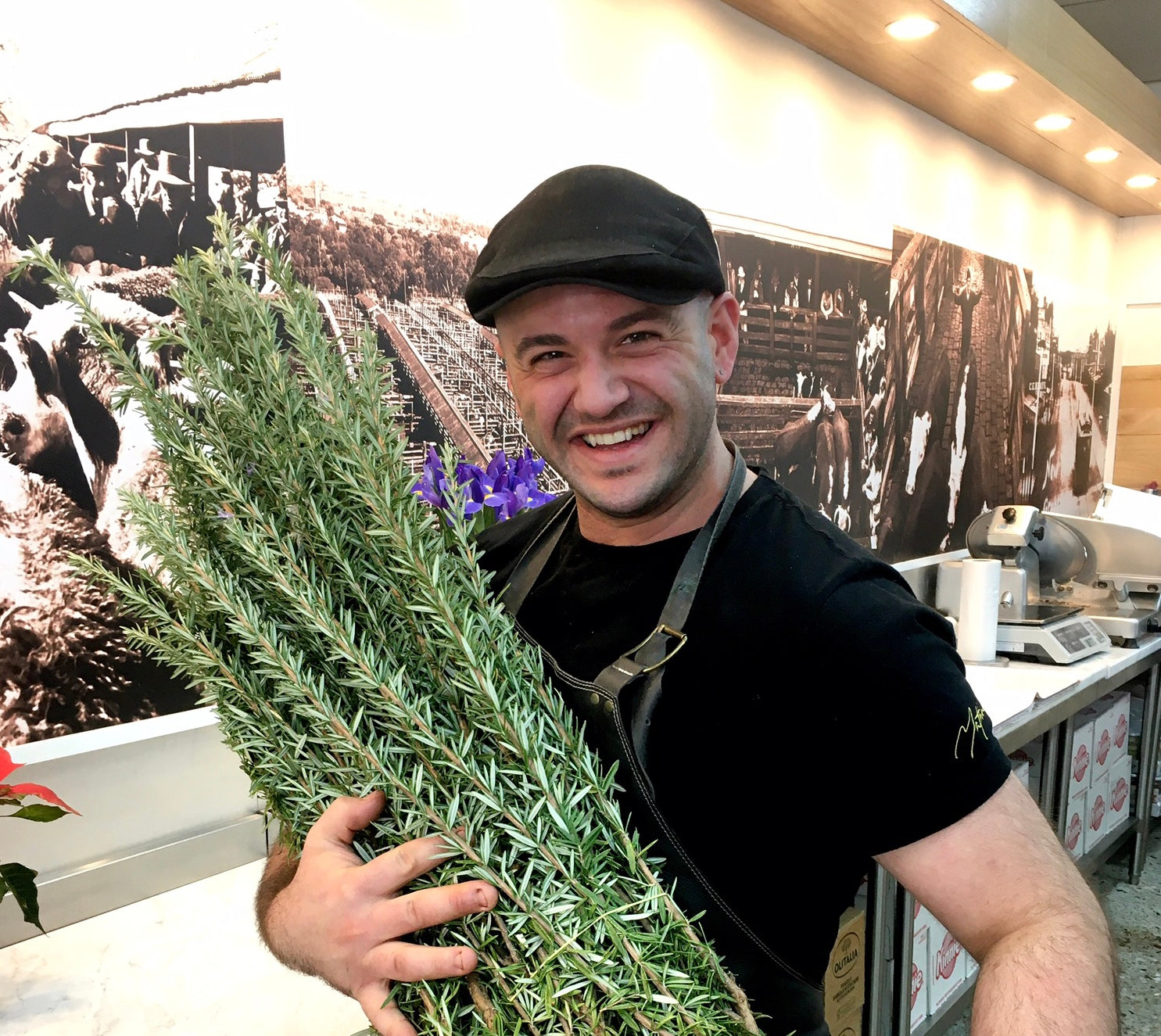 Mathew Bonnici in his flat cap and apron, holding an armful of fresh rosemary in the shop